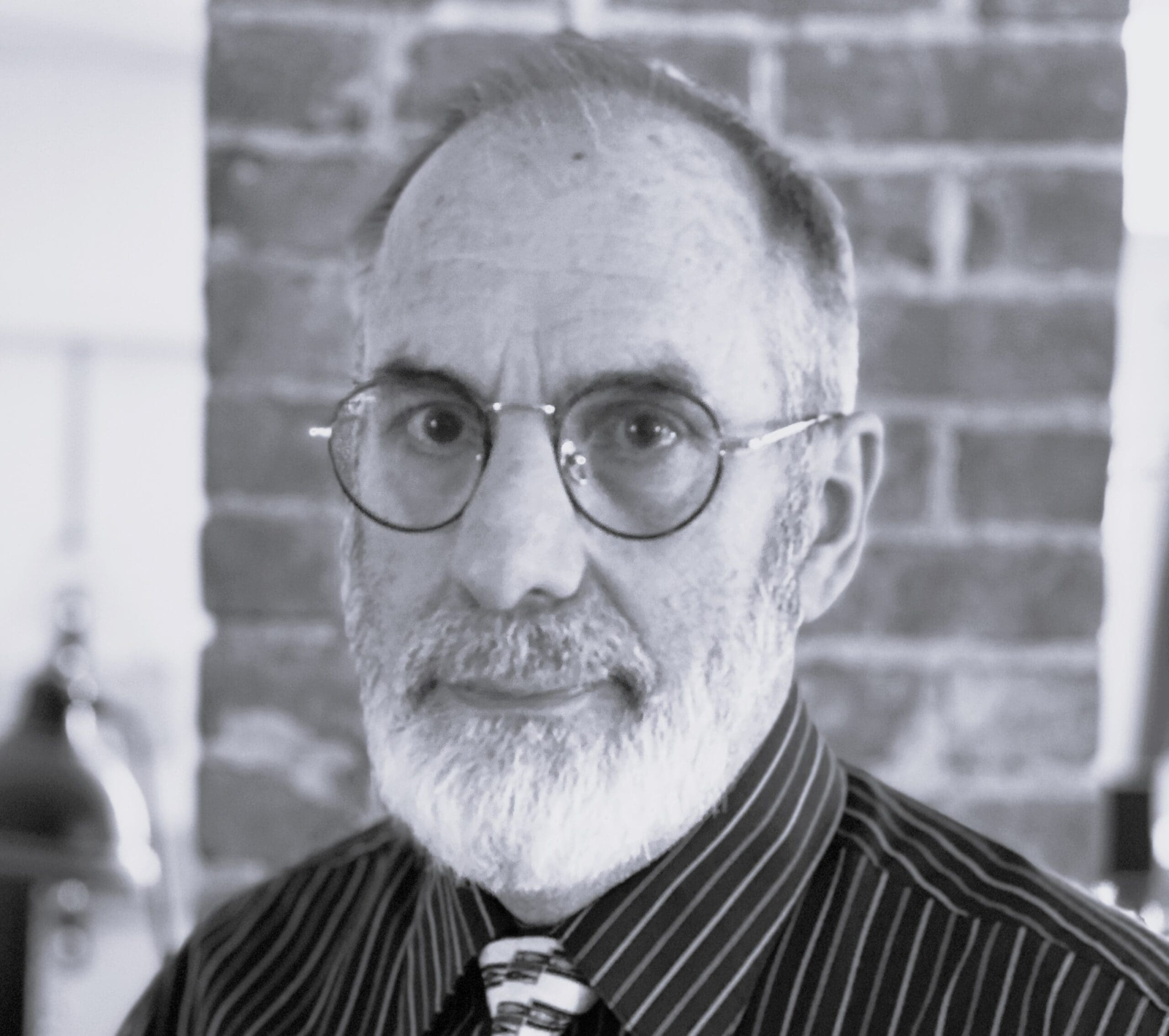 A person with glasses and a beard in front of a brick wall.