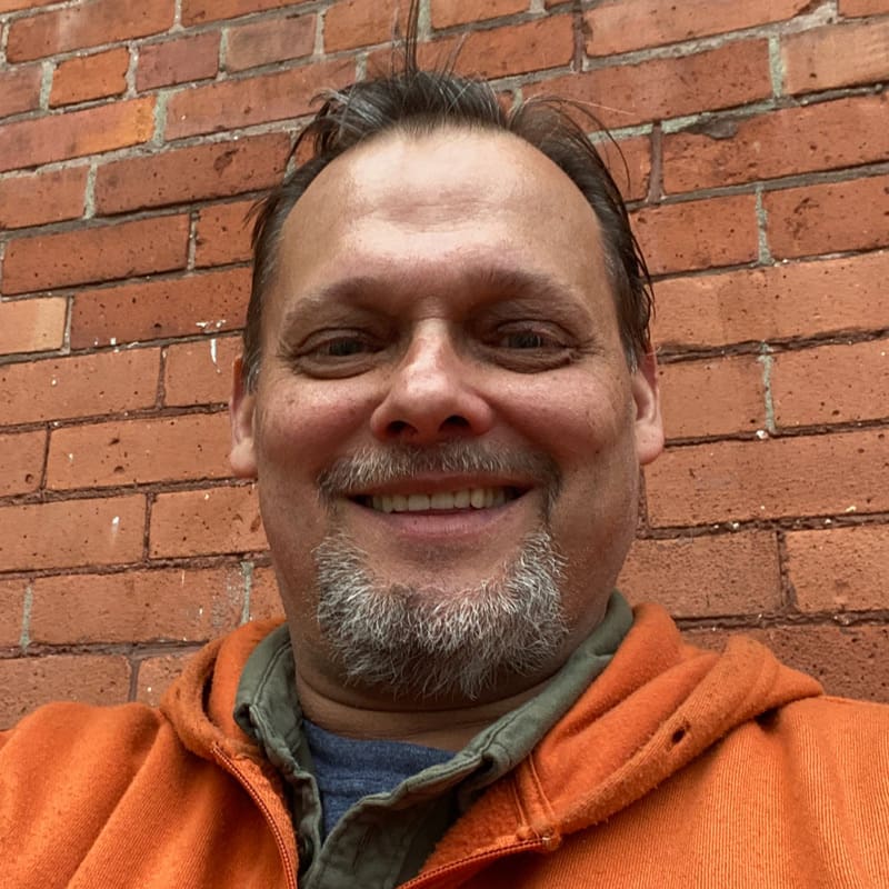 Smiling person in front of a brick wall wearing an orange jacket.