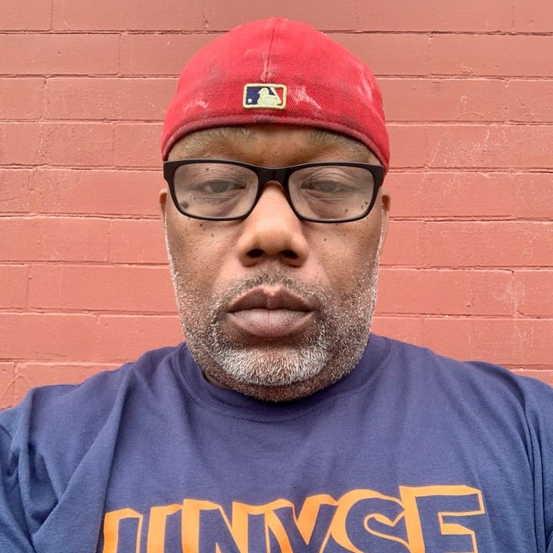 Person in red cap and glasses standing against a brick wall.