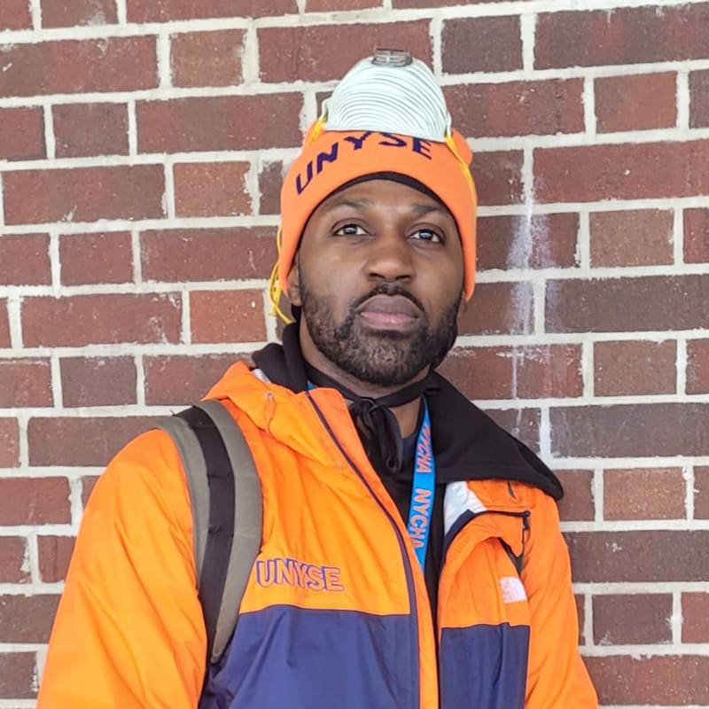Man in orange UNYSE hat and jacket, standing against a brick wall.
